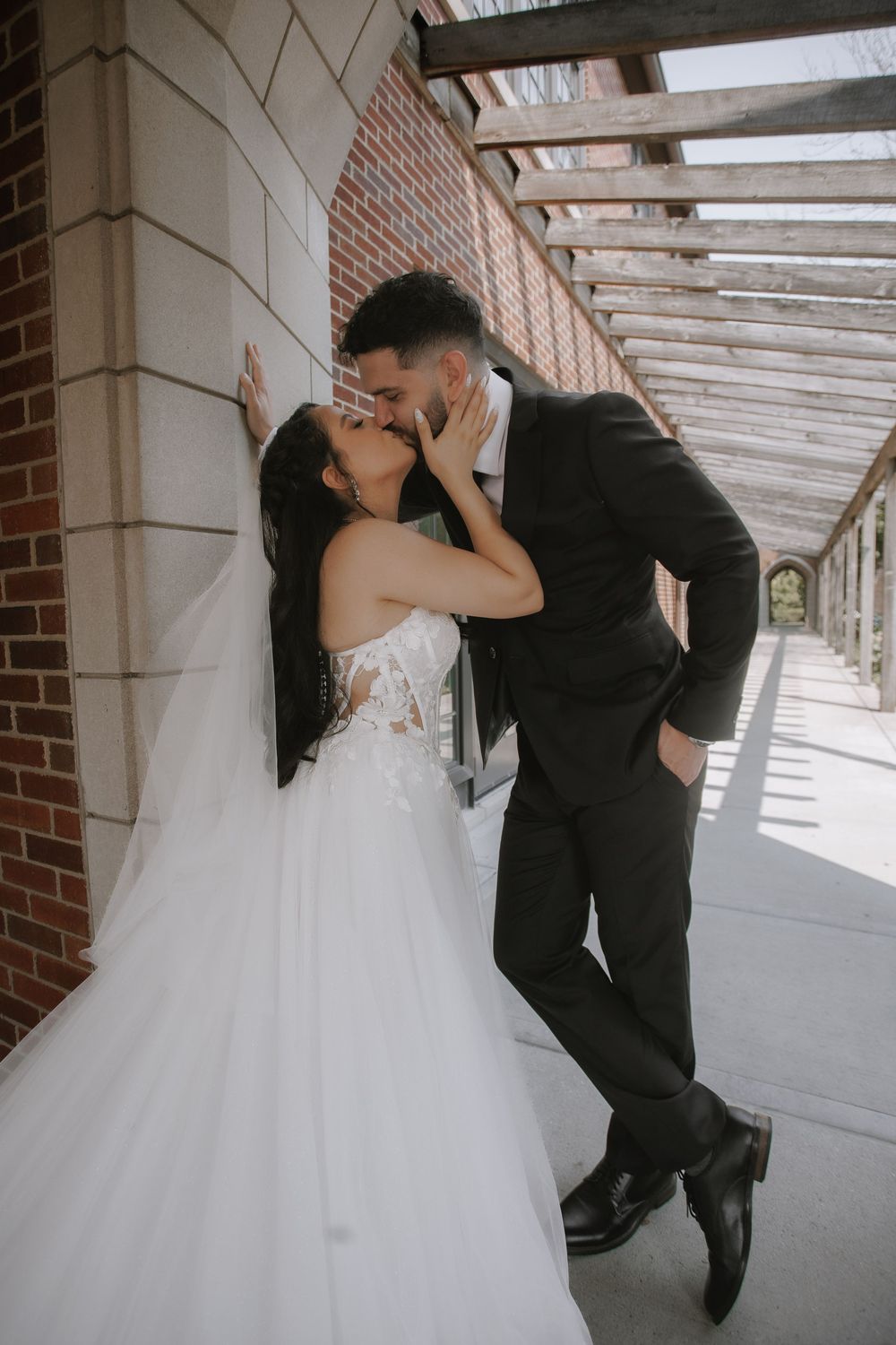 A romantic wedding couple shares five intimate kisses while standing in a covered outdoor walkway with brick pillars.