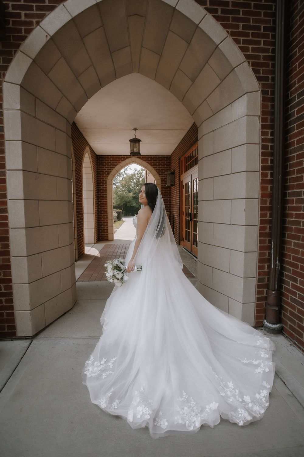 A flowing white wedding gown with intricate lace details trails elegantly through a brick and stone archway corridor.
