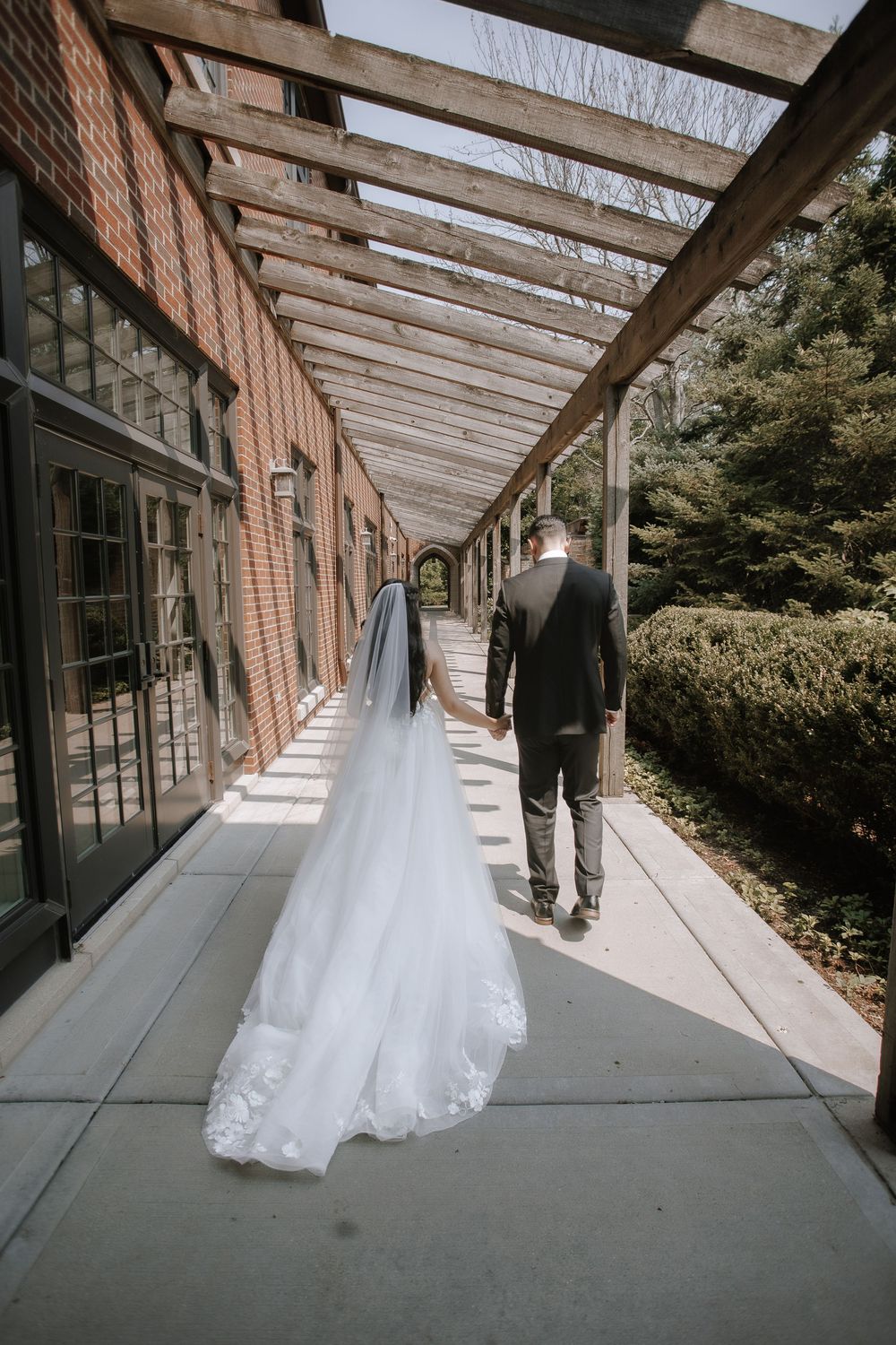 A wedding dress train trails behind as a couple walks down a covered brick colonnade walkway.