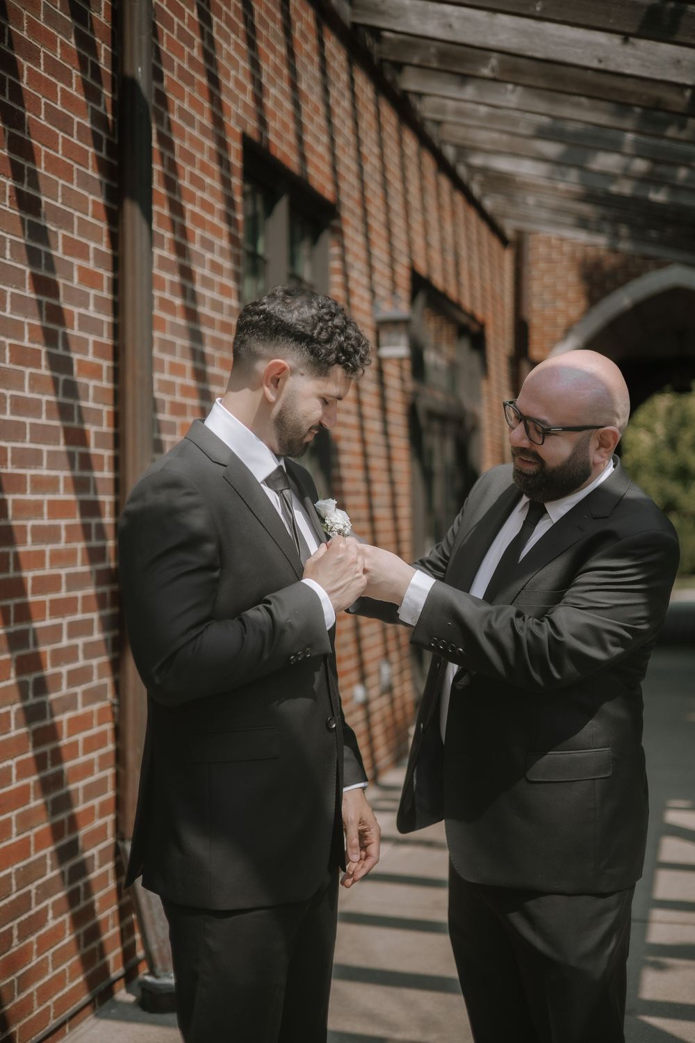Wedding party members in black suits share a lighthearted moment together against a brick wall backdrop.