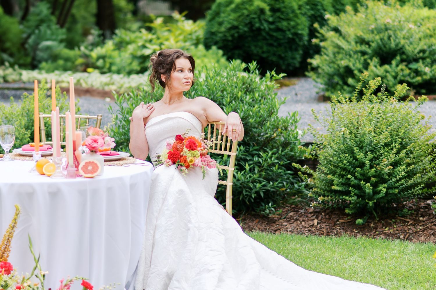 A couple in formal attire poses in a garden setting with a flowing pink tulle gown spread across the grass.