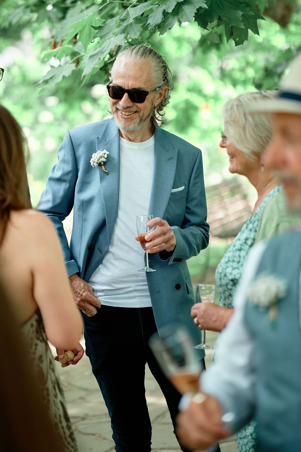 People enjoying drinks and conversation at an outdoor garden party in a casual yet elegant setting, with greenery in background.