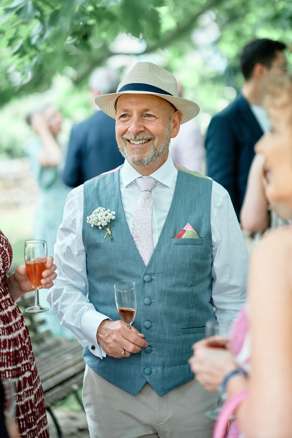 A guest in a gray vest and white shirt smiles while holding a beverage at an outdoor garden party.