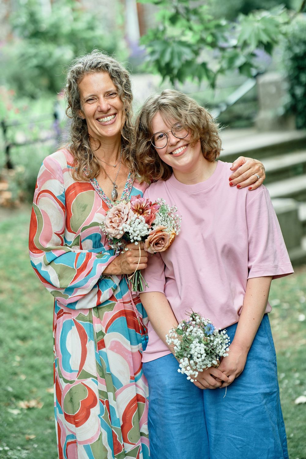 Two people in casual attire share a tender moment outdoors while holding flowers in a lush green garden setting.