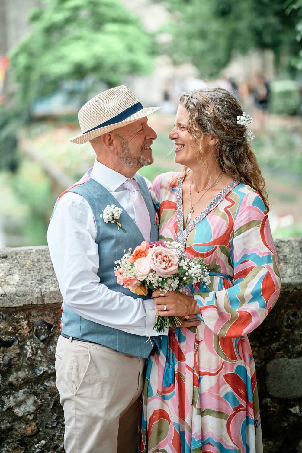 Happy mature couple embraces and laughs outdoors in summer garden while wearing colorful attire and floral bouquet.