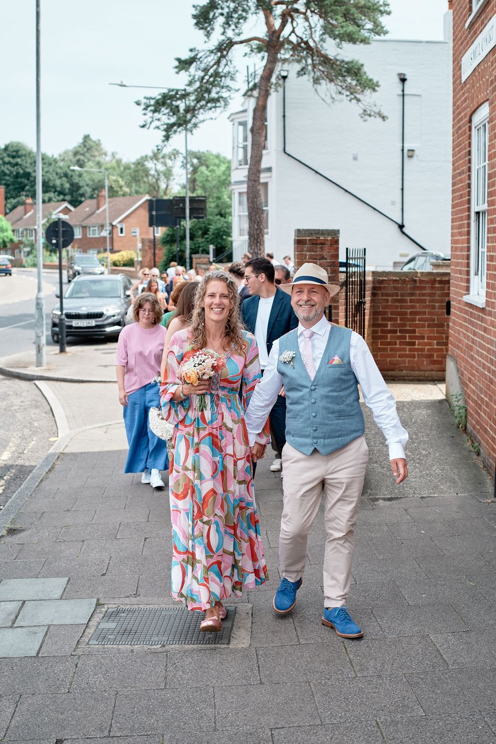 A couple walks together down a paved sidewalk outside brick buildings on a sunny summer day.