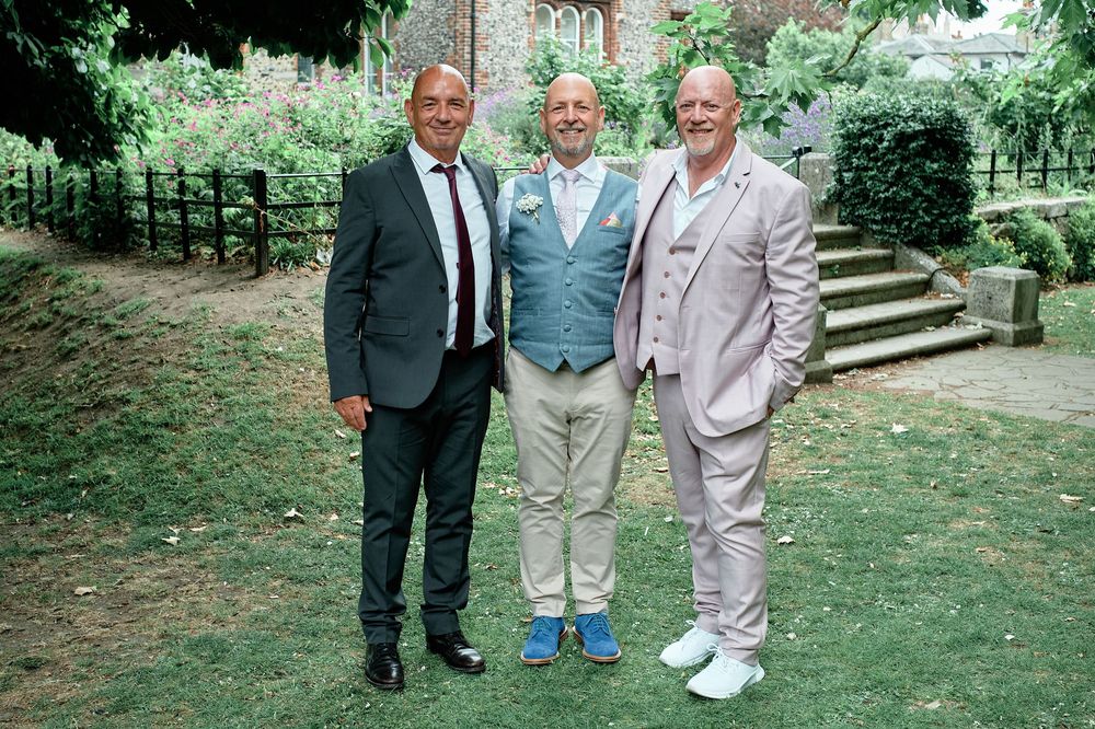Three people in formal attire stand together on a grassy lawn near a brick building in the background.