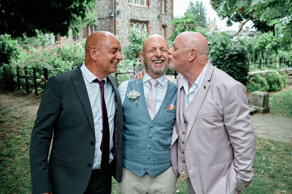 Three people engaging in cheerful conversation while standing in a lush garden setting at a formal event.