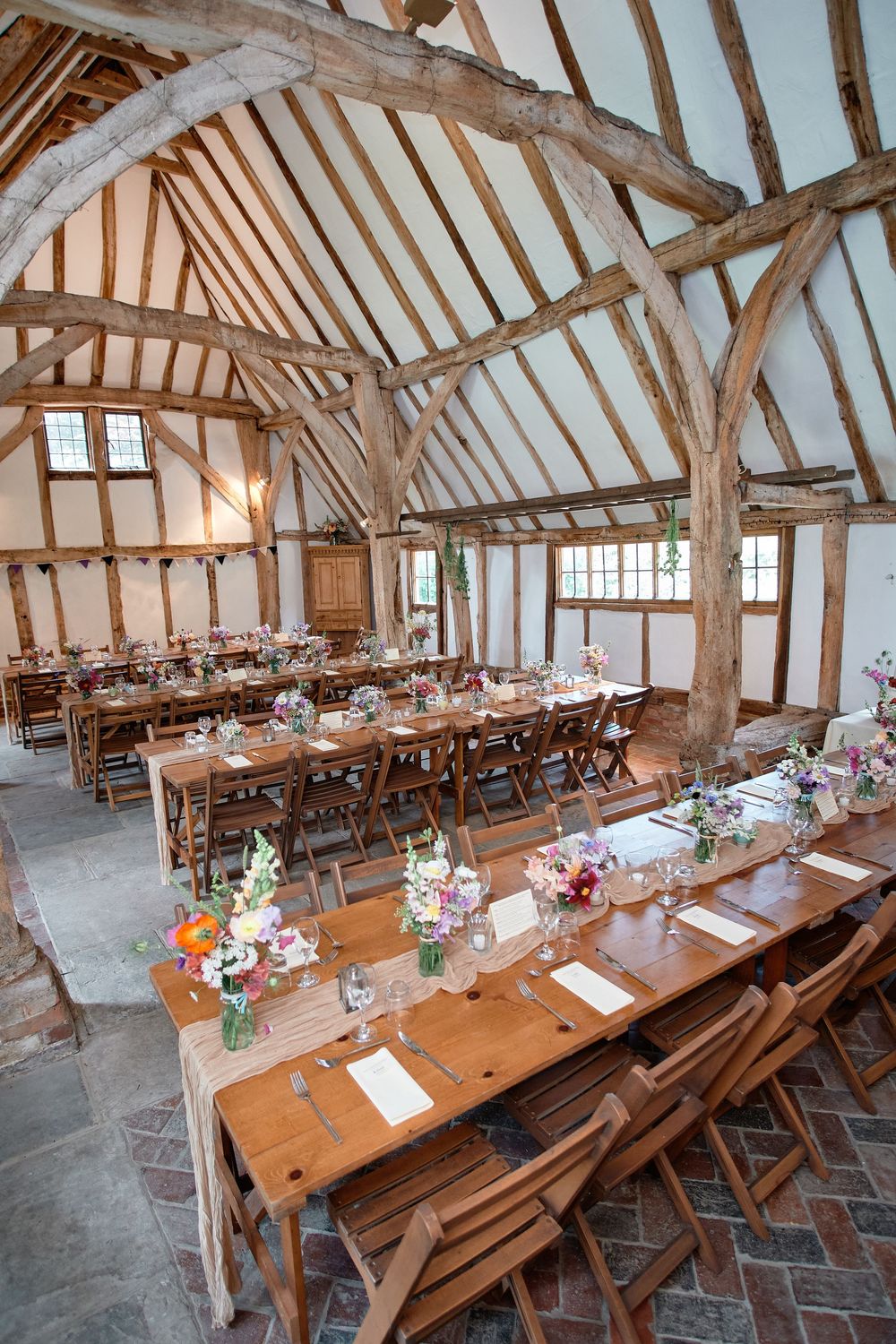 Rustic wooden barn venue with long farmhouse tables decorated with floral centerpieces and cross-back chairs.