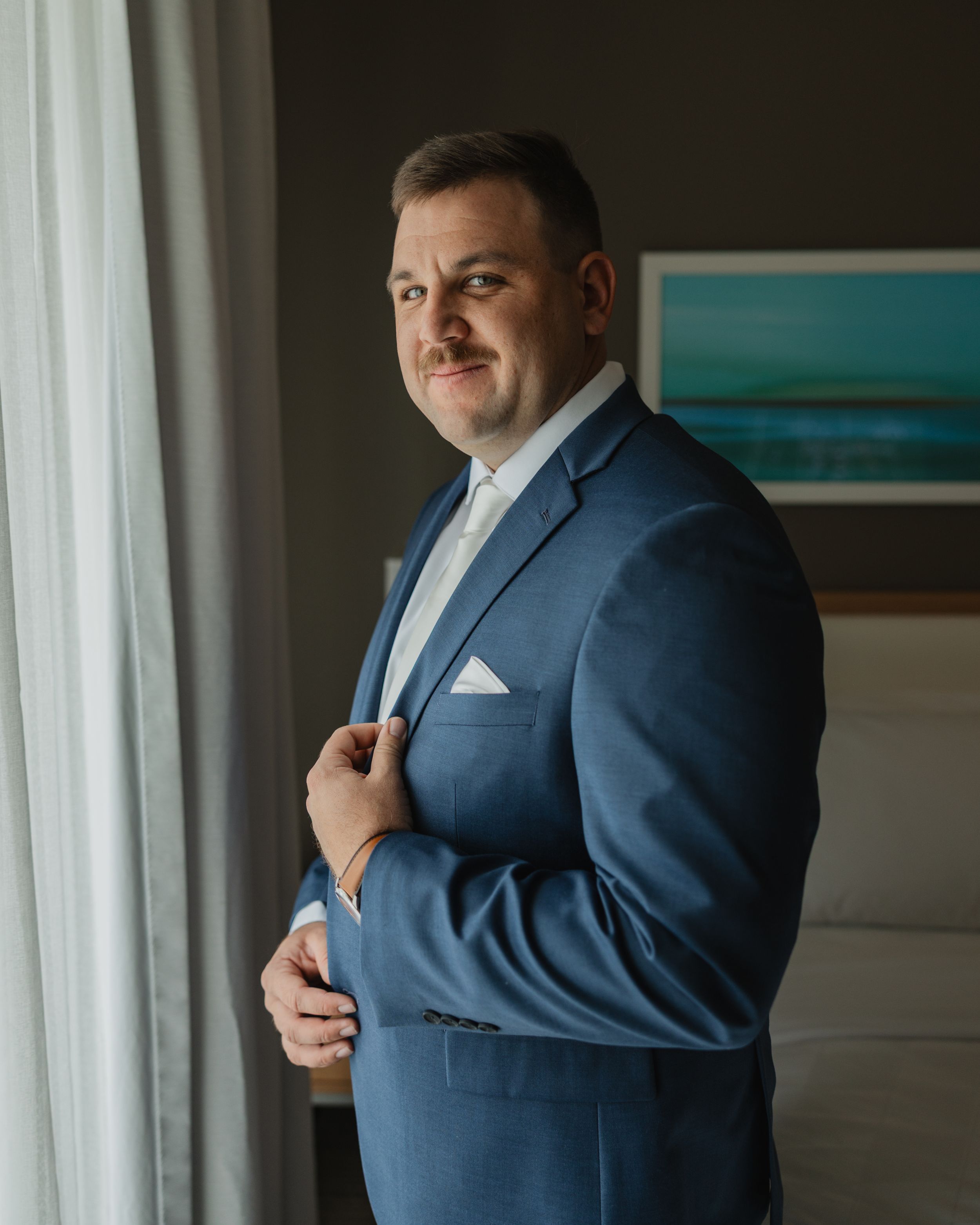 Professional portrait of a man in a blue suit and white pocket square standing by a window in natural light.