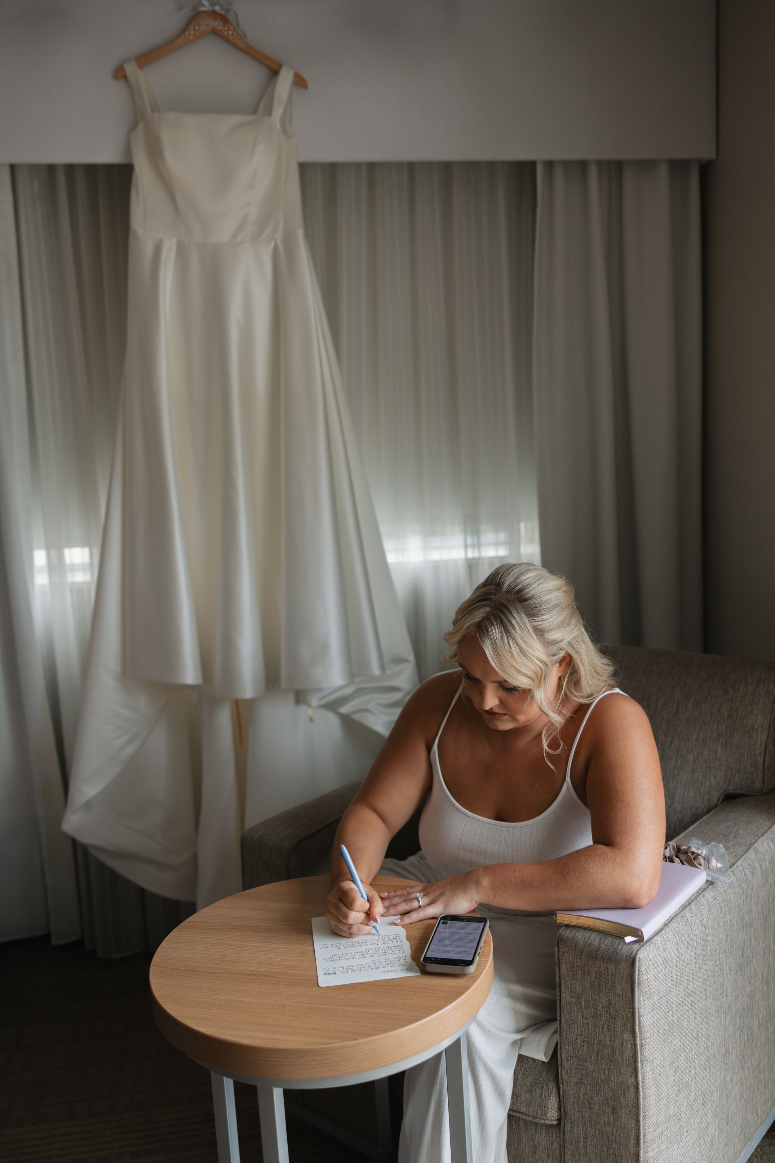 Wedding dress hangs above a person writing at a round wooden table in a softly lit room.