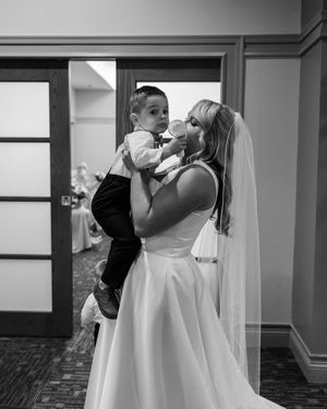 A tender black and white moment shows a bride in a flowing dress and veil holding a small child during a wedding celebration.