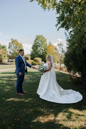 A scenic outdoor wedding photo showing a couple in formal attire standing on a grassy lawn with trees in the background.
