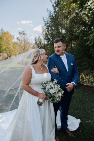 Wedding couple posing outdoors with white bouquet and flowing veil against autumn trees.