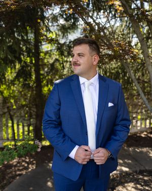 Groom in navy blue suit and white shirt poses in a garden setting during a wedding photoshoot.