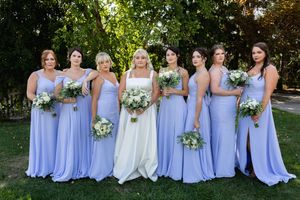 A wedding party poses outdoors with bridesmaids in matching periwinkle dresses holding floral bouquets.