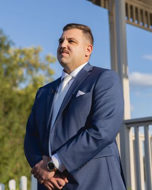 Person in a navy blue suit standing on an outdoor porch with white columns and trees in background.