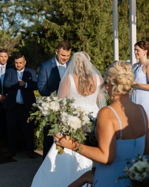 Wedding guests gather outdoors for a ceremony under trees with sunlight filtering through the branches.