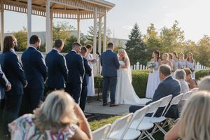 Wedding guests watch ceremony under white gazebo at outdoor venue during sunset.