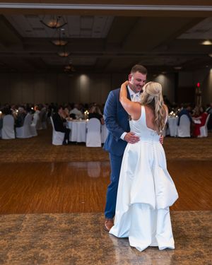 Wedding couple shares romantic first dance in dimly lit ballroom with wooden floors and white chair covers.