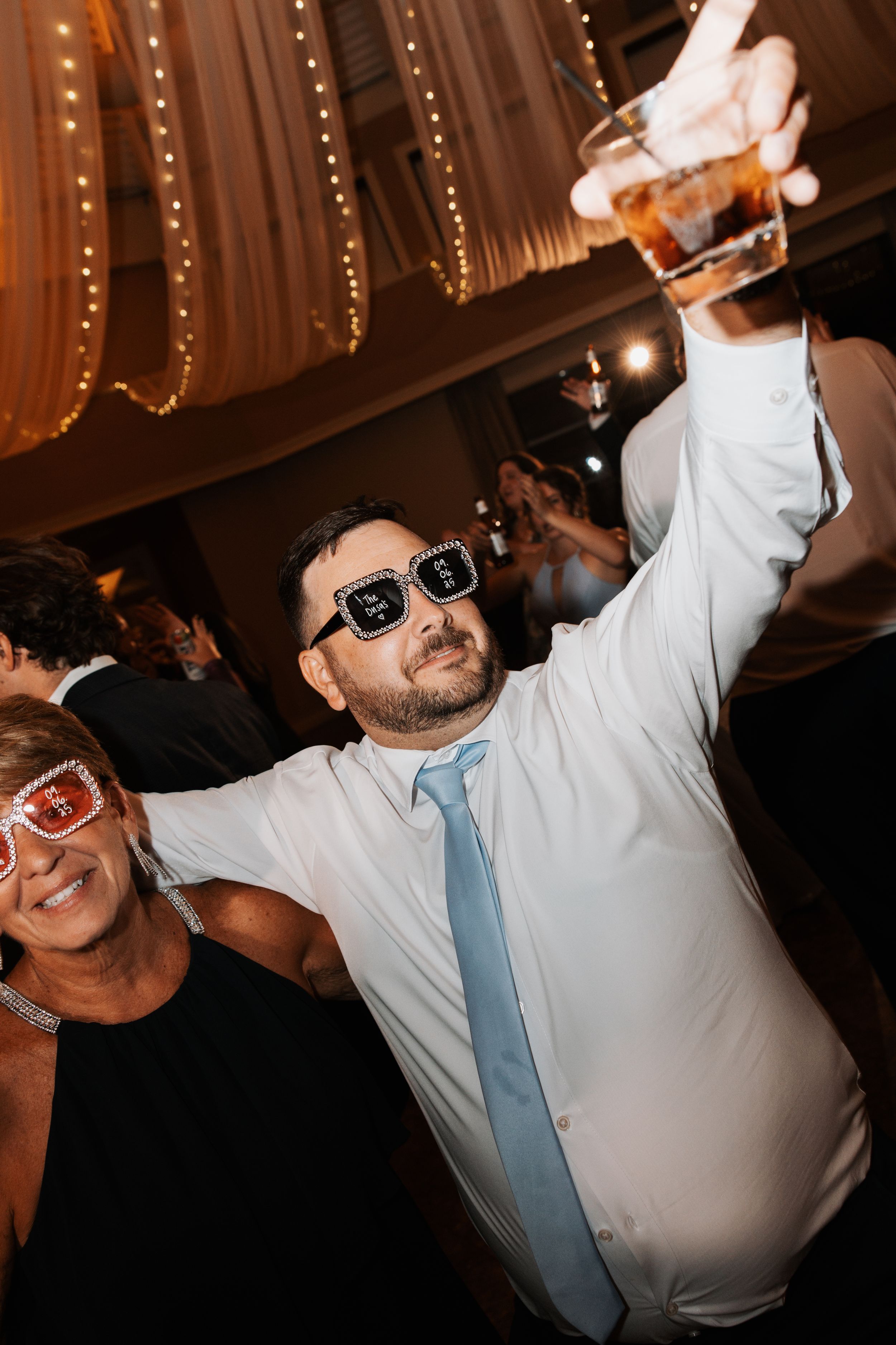 Wedding guests in sunglasses celebrate enthusiastically on the dance floor with string lights illuminating the venue.