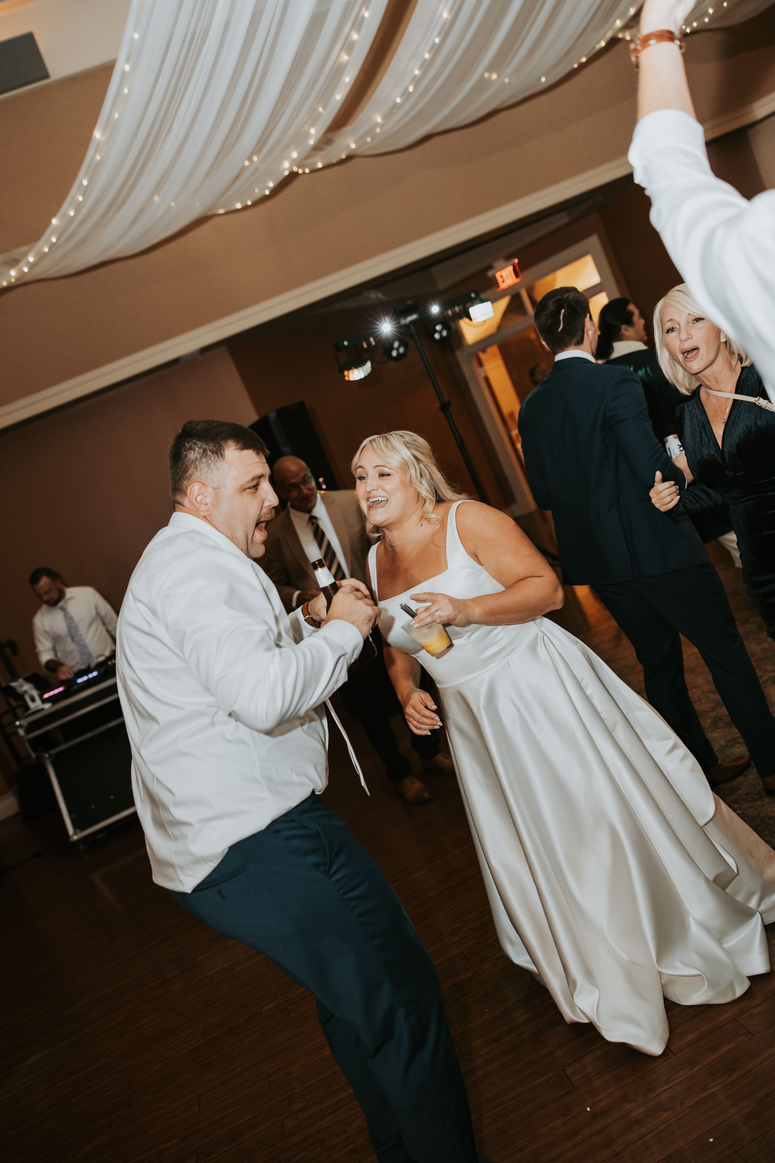 Couple dances energetically at wedding reception while other guests mingle on dance floor under string lights.