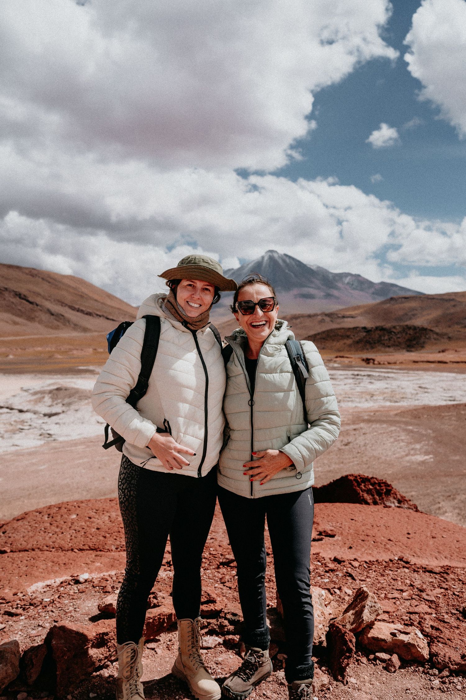 Two hikers pose together on rocky terrain with mountain peaks in background.