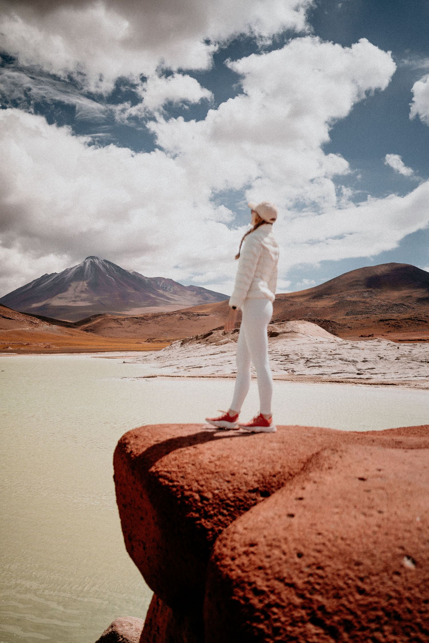 Person in white stands on red rock overlooking salt flats with mountains in background.