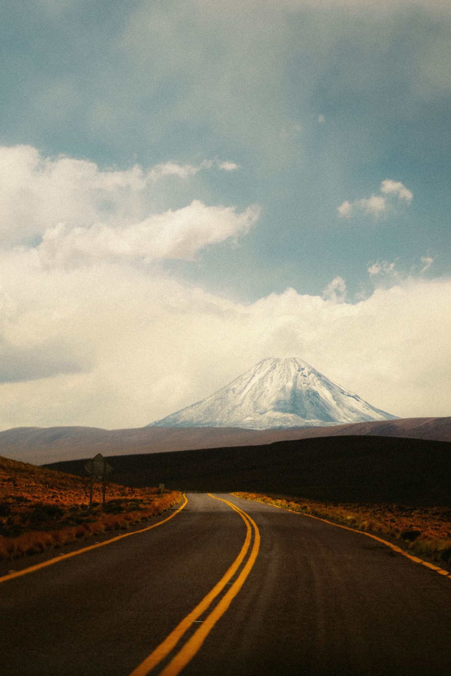 Empty desert highway leading towards snow-capped mountains under a cloudy blue sky.