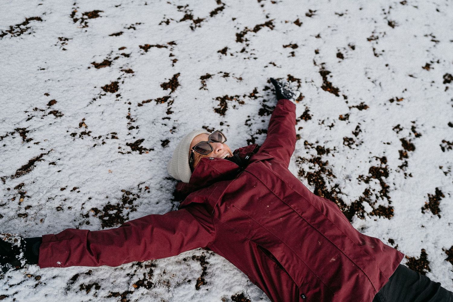 Person in burgundy jacket lying playfully in the snow against mountain backdrop.