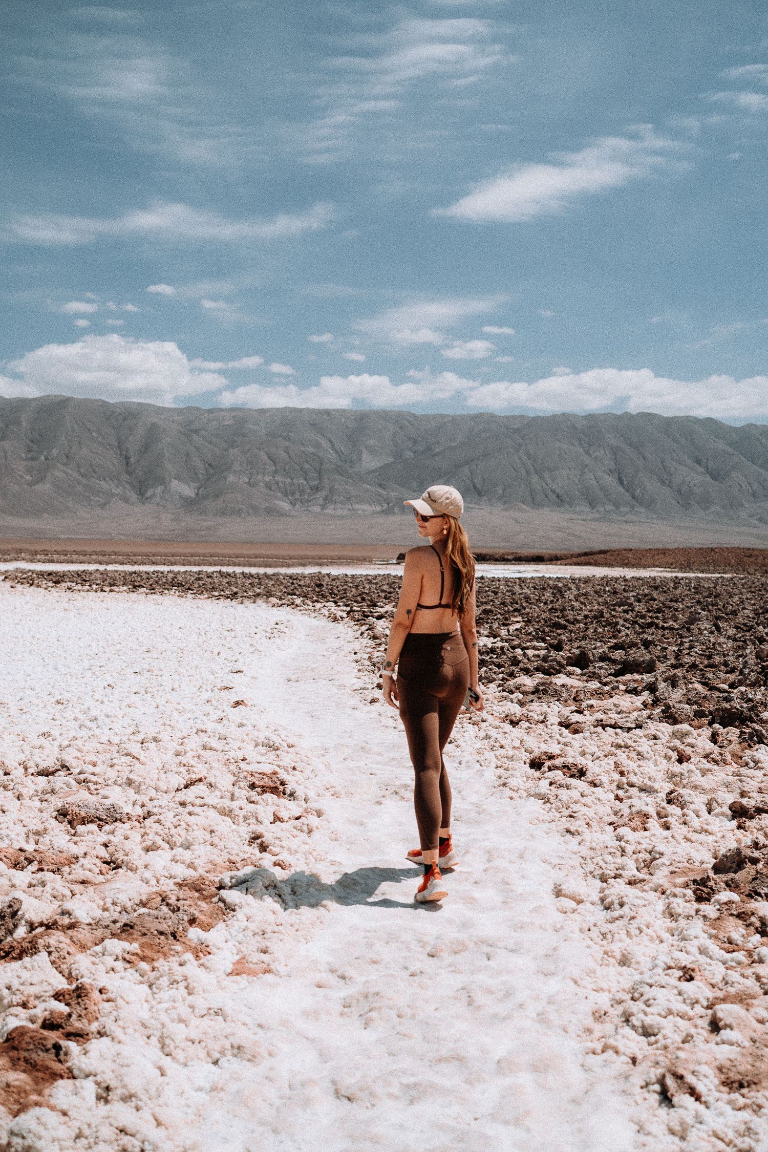 A lone traveler in brown attire walks along a salt flat path surrounded by rugged mountains under a blue sky.