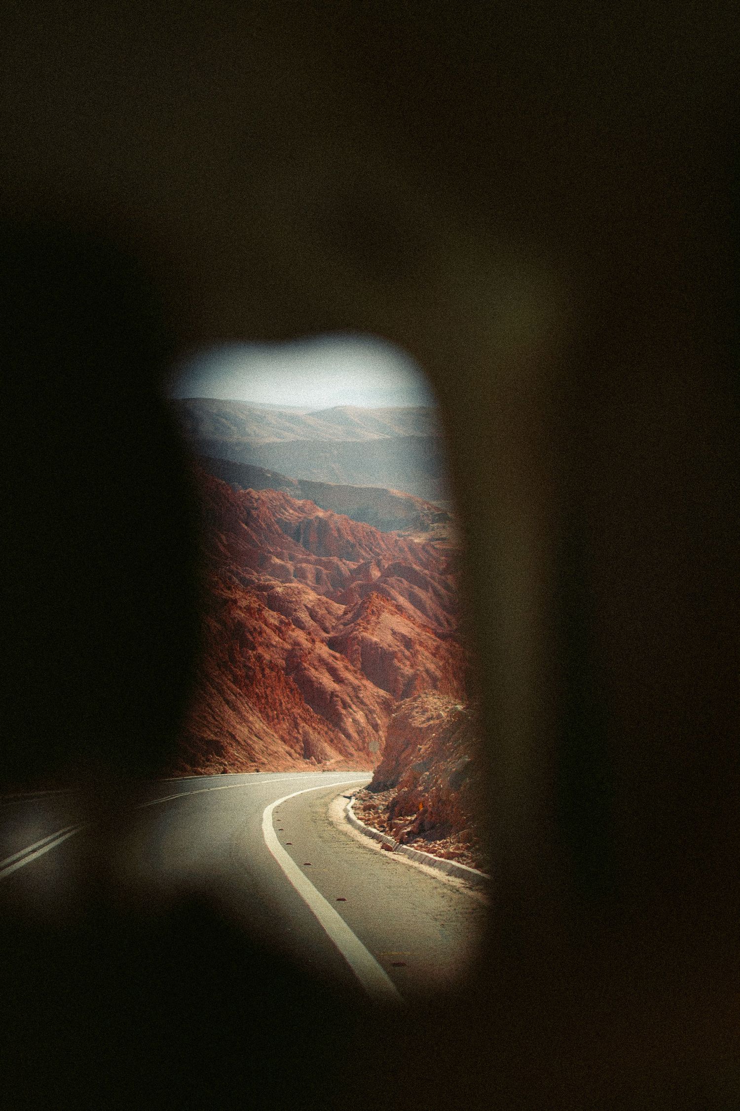 A winding mountain road viewed through a car window with dramatic red rock formations in the distance.