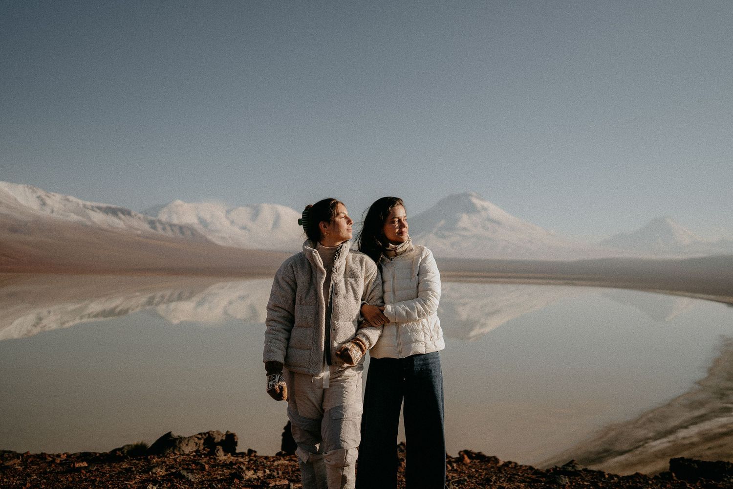 Two people standing by a reflective lake with snow-capped mountains in the background.