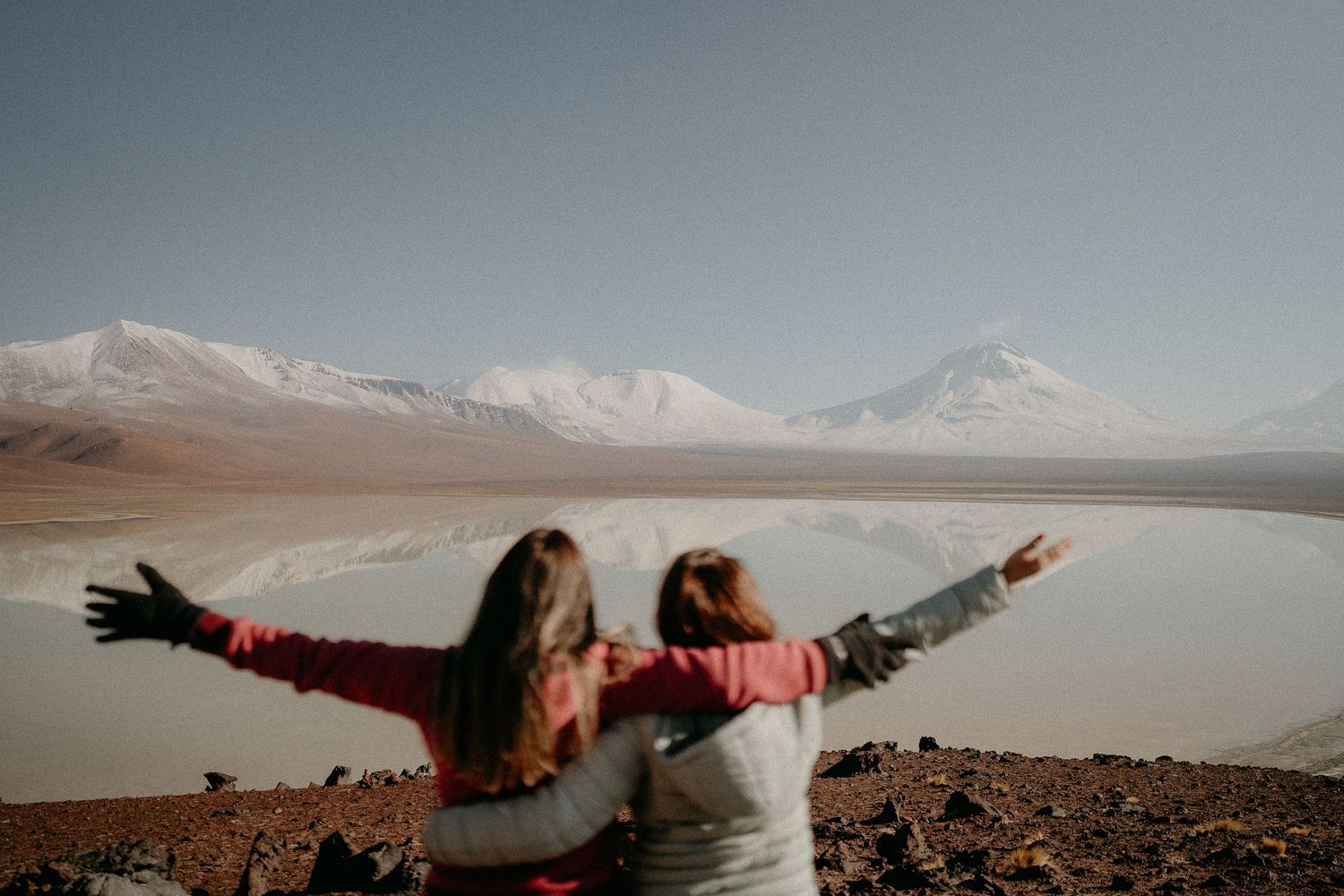 Two friends with outstretched arms enjoying mountain views by a reflective lake.
