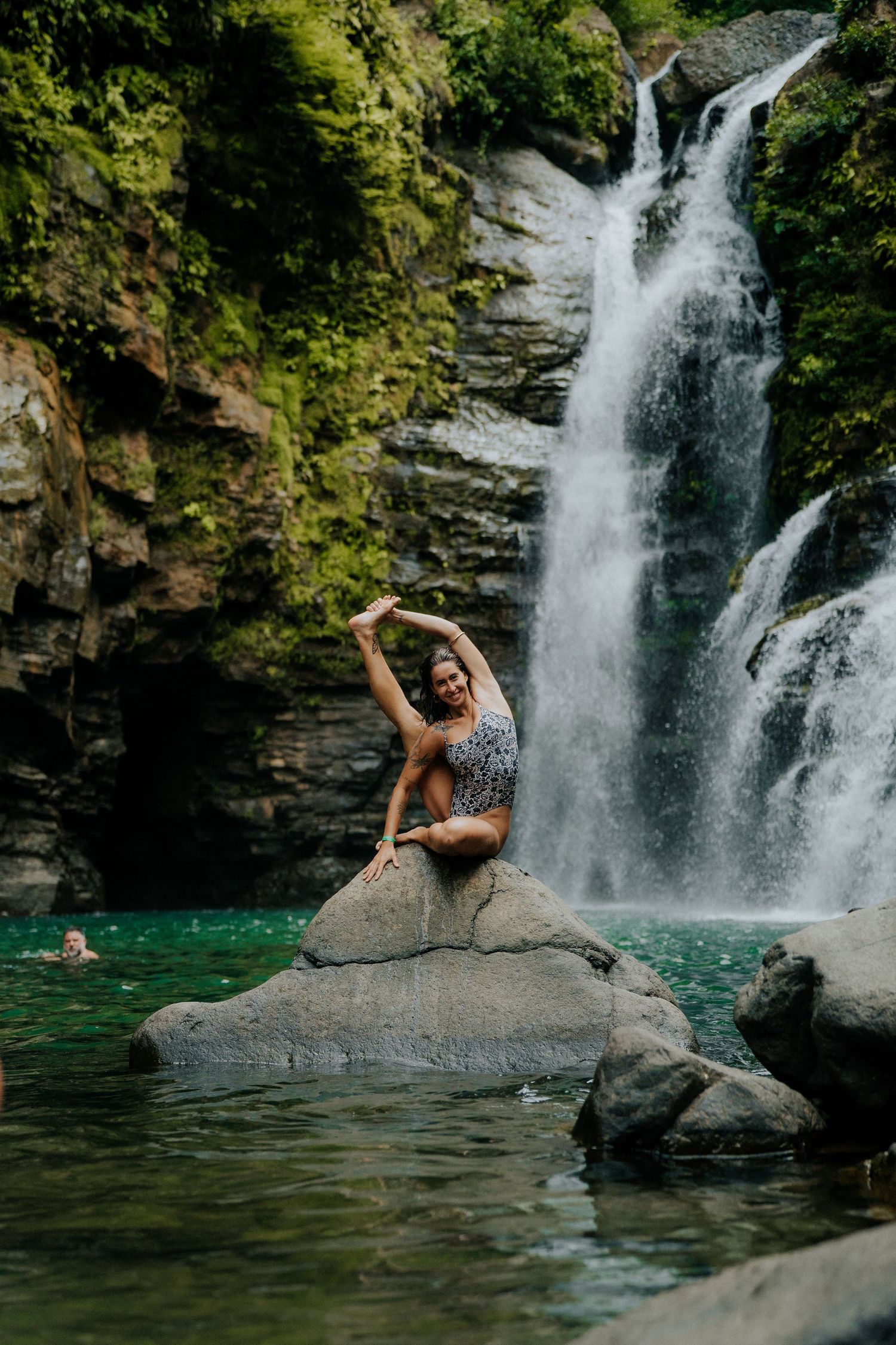 Person posing artistically on rock near dramatic waterfall and swimming hole.