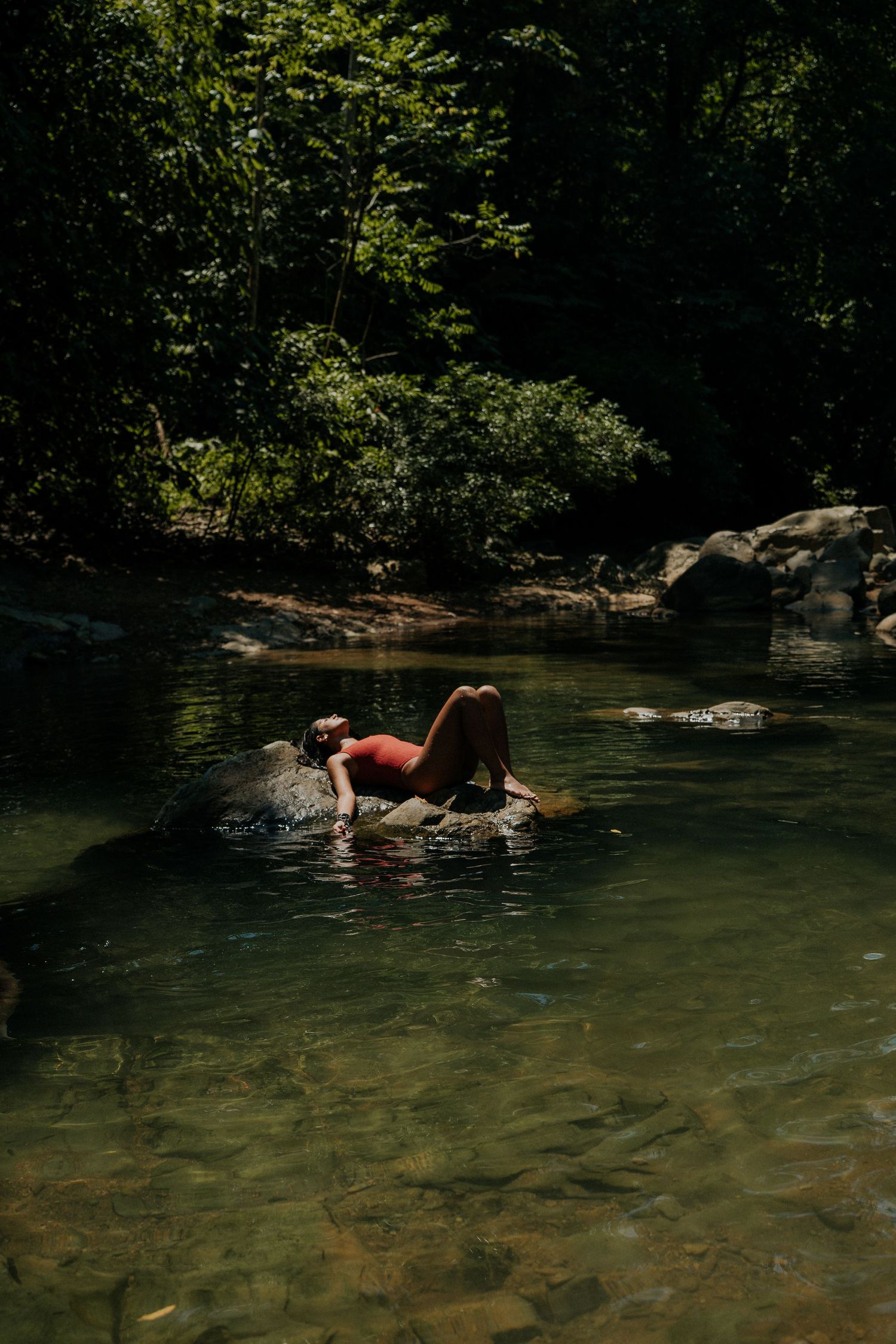 Someone relaxes on a rock in a serene forest swimming hole surrounded by lush greenery.