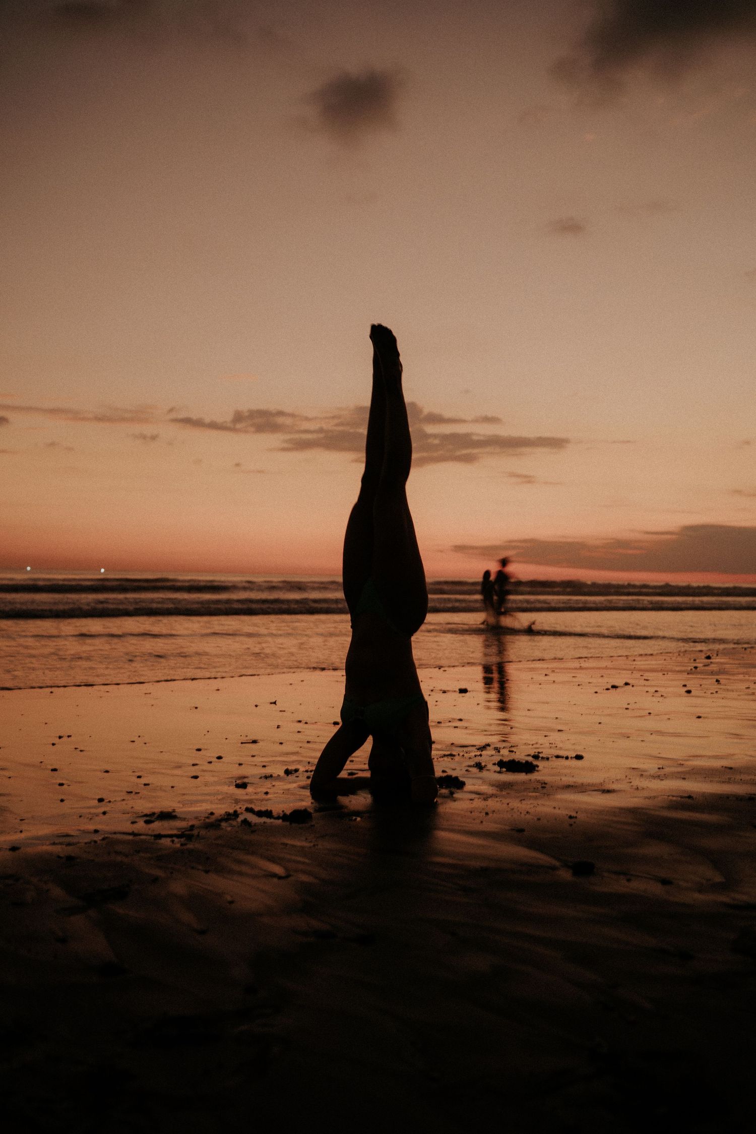 Silhouetted figure performs a handstand on a beach at sunset with another person visible in the distance.