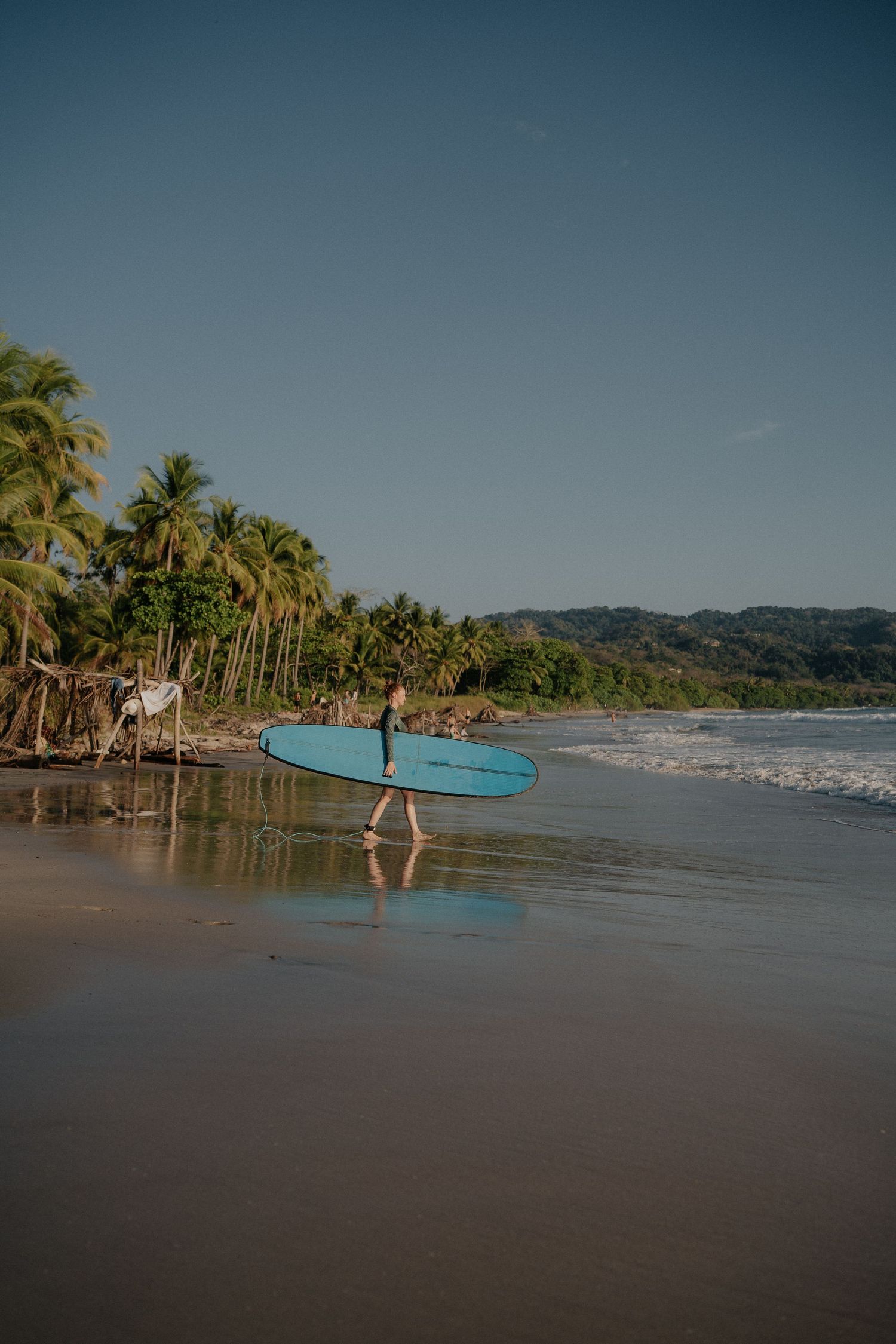 Surfer carrying blue surfboard along tropical beach with palm trees and mountains in background.