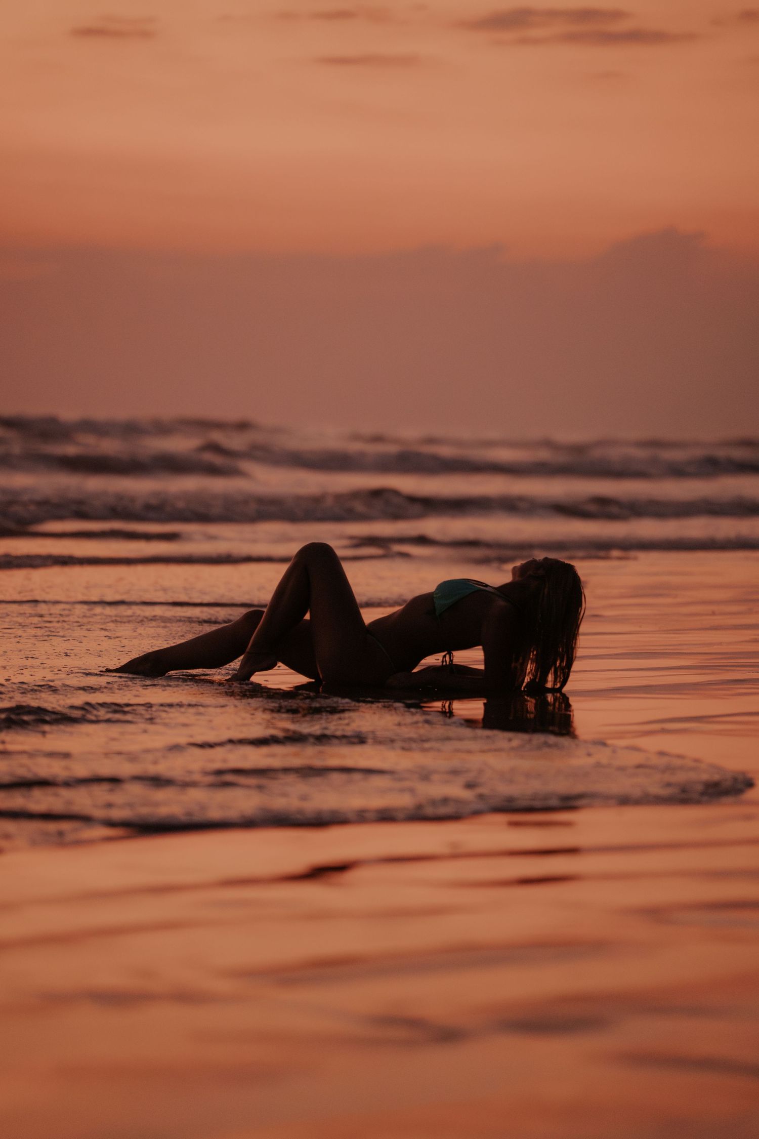 Surfer silhouette floating in ocean waves during dramatic orange sunset.