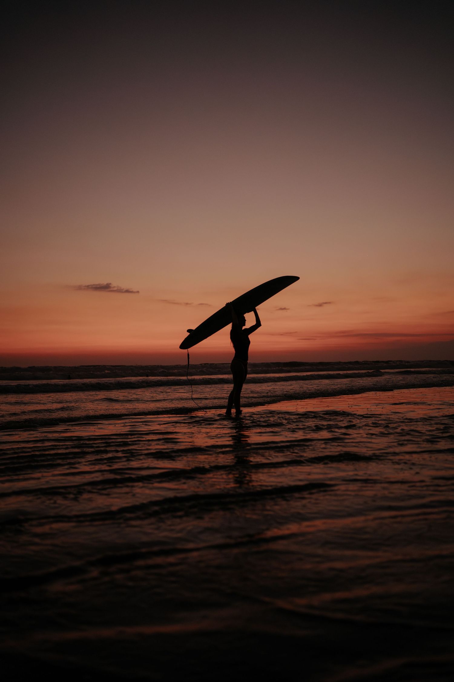 Silhouette of surfer carrying board against dramatic red sunset sky.