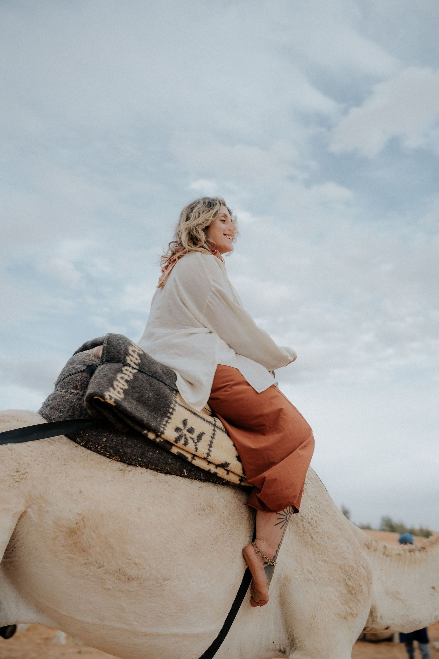Desert traveler rides atop a decorated camel through sandy terrain under cloudy skies.