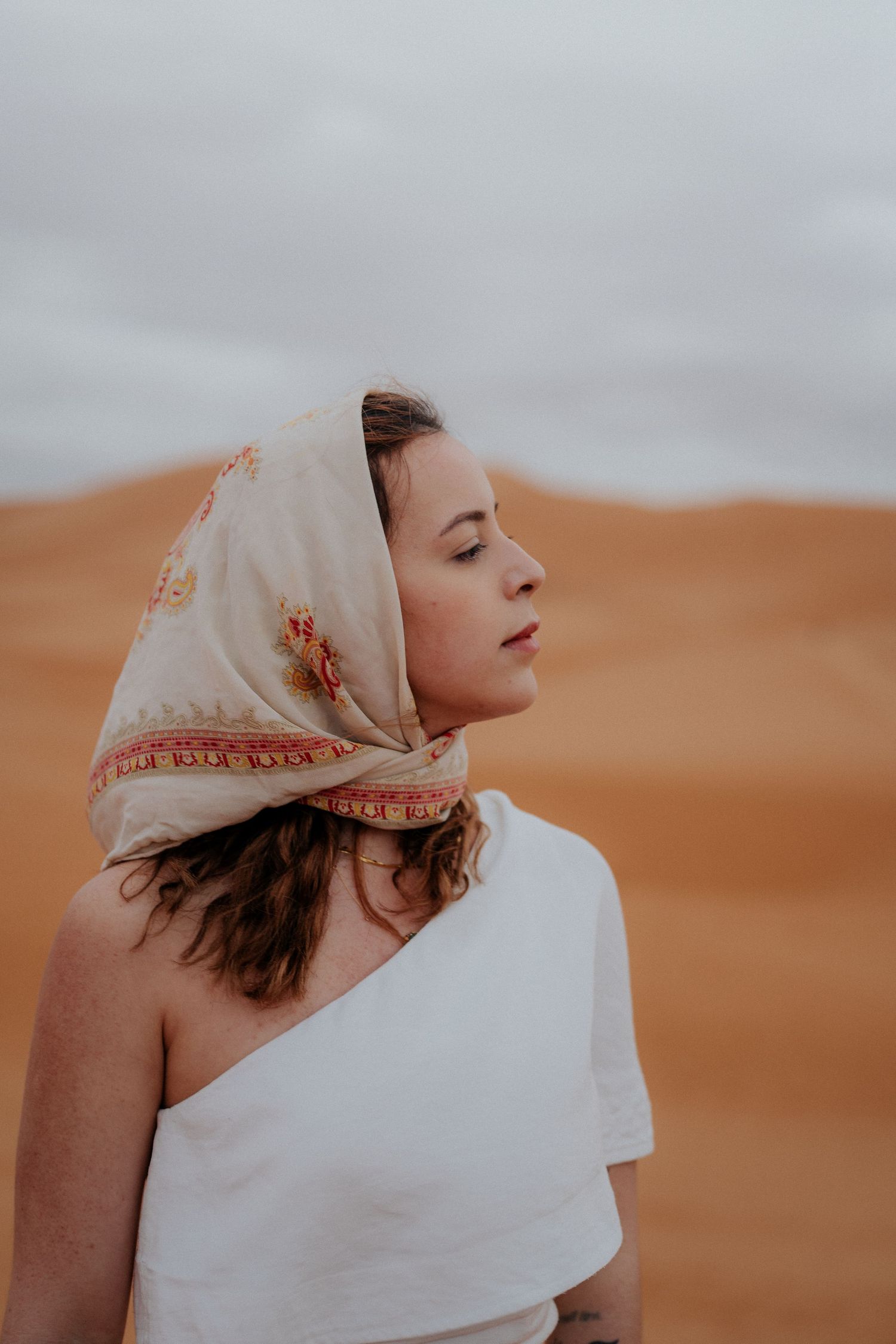 Profile view against desert dunes shows flowing fabric and traditional head covering in warm light.