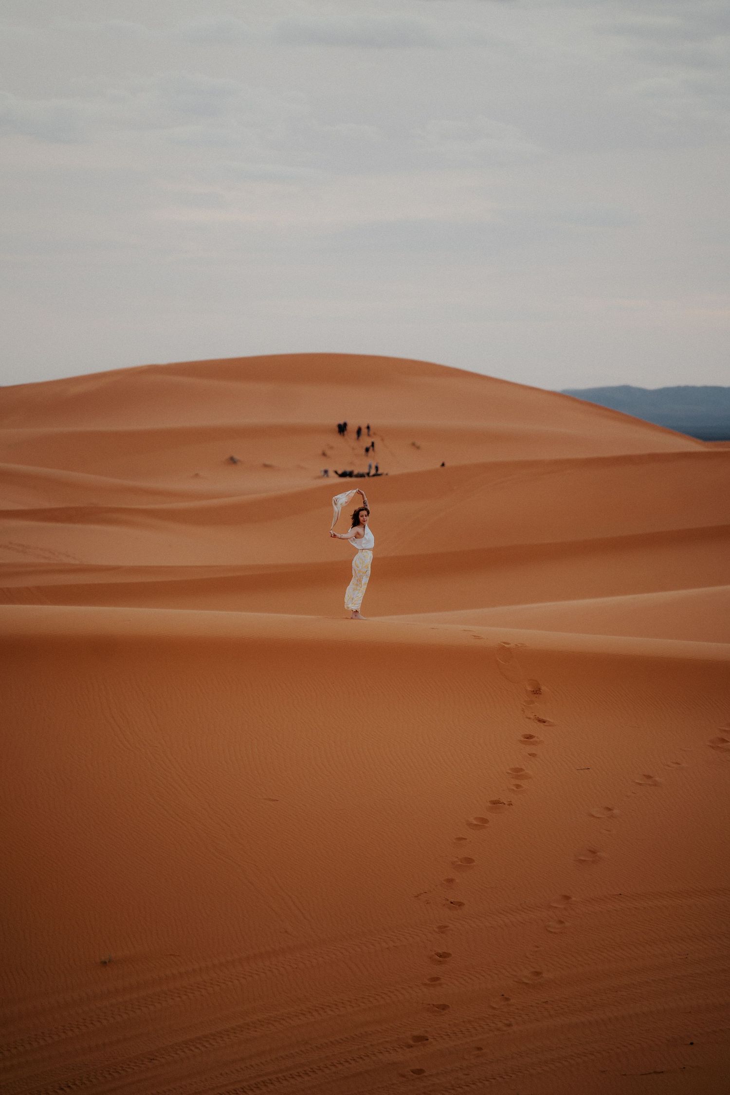 Distant figure walks along the ridge of massive orange sand dunes leaving footprints behind.