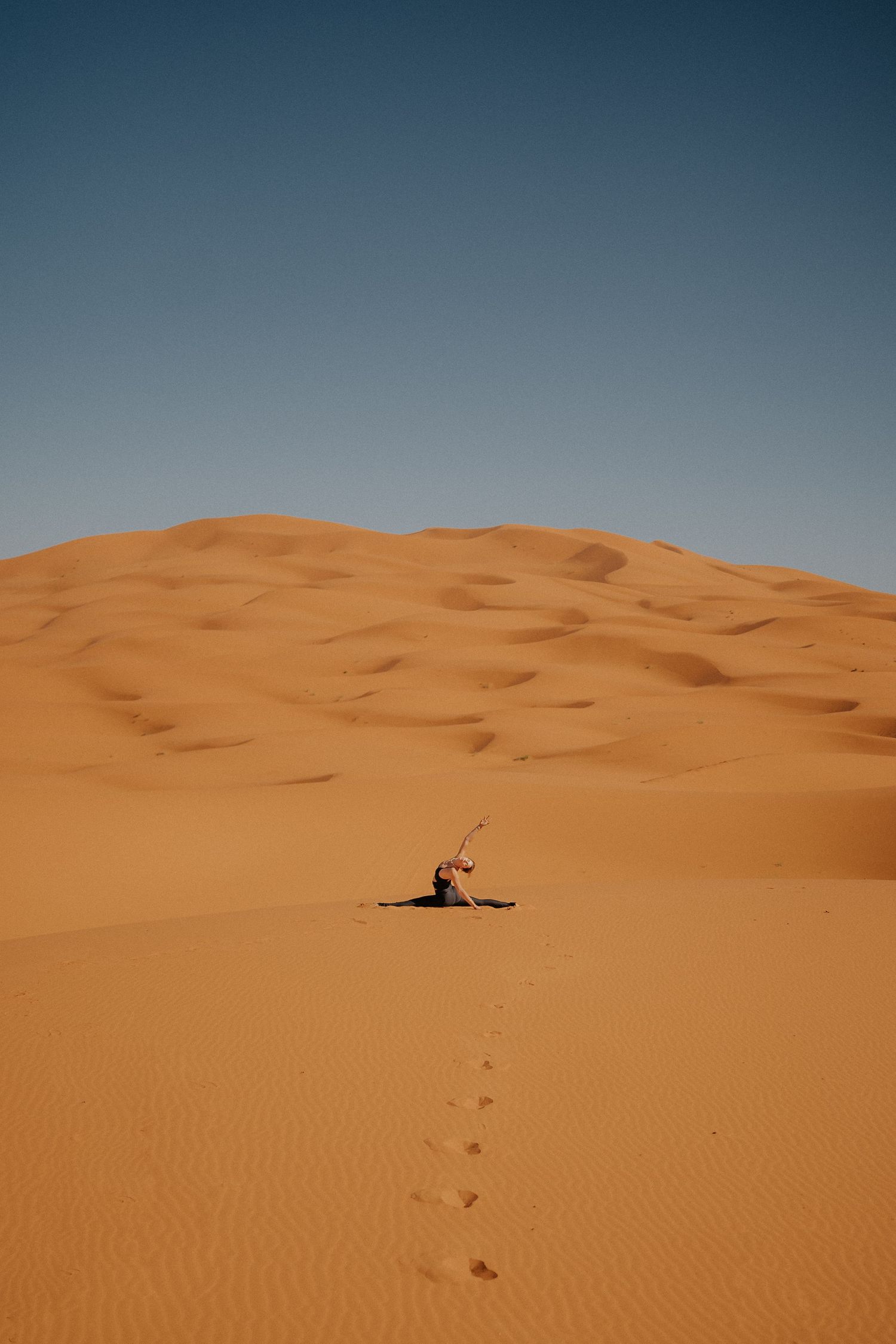 Minimalist desert landscape shows tiny figure sitting among vast orange sand dunes.