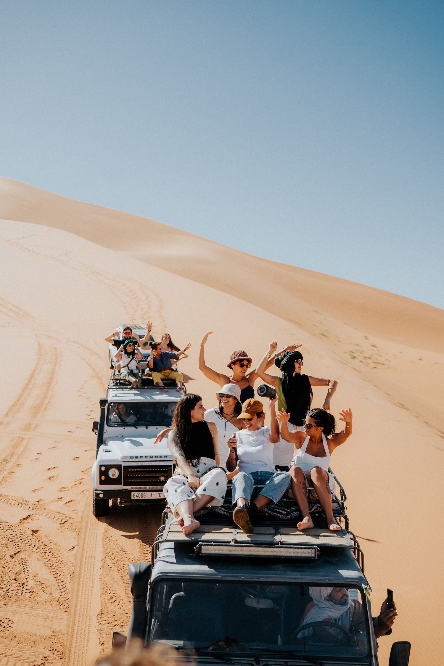 Group riding on top of off-road vehicles traversing desert sand dunes.