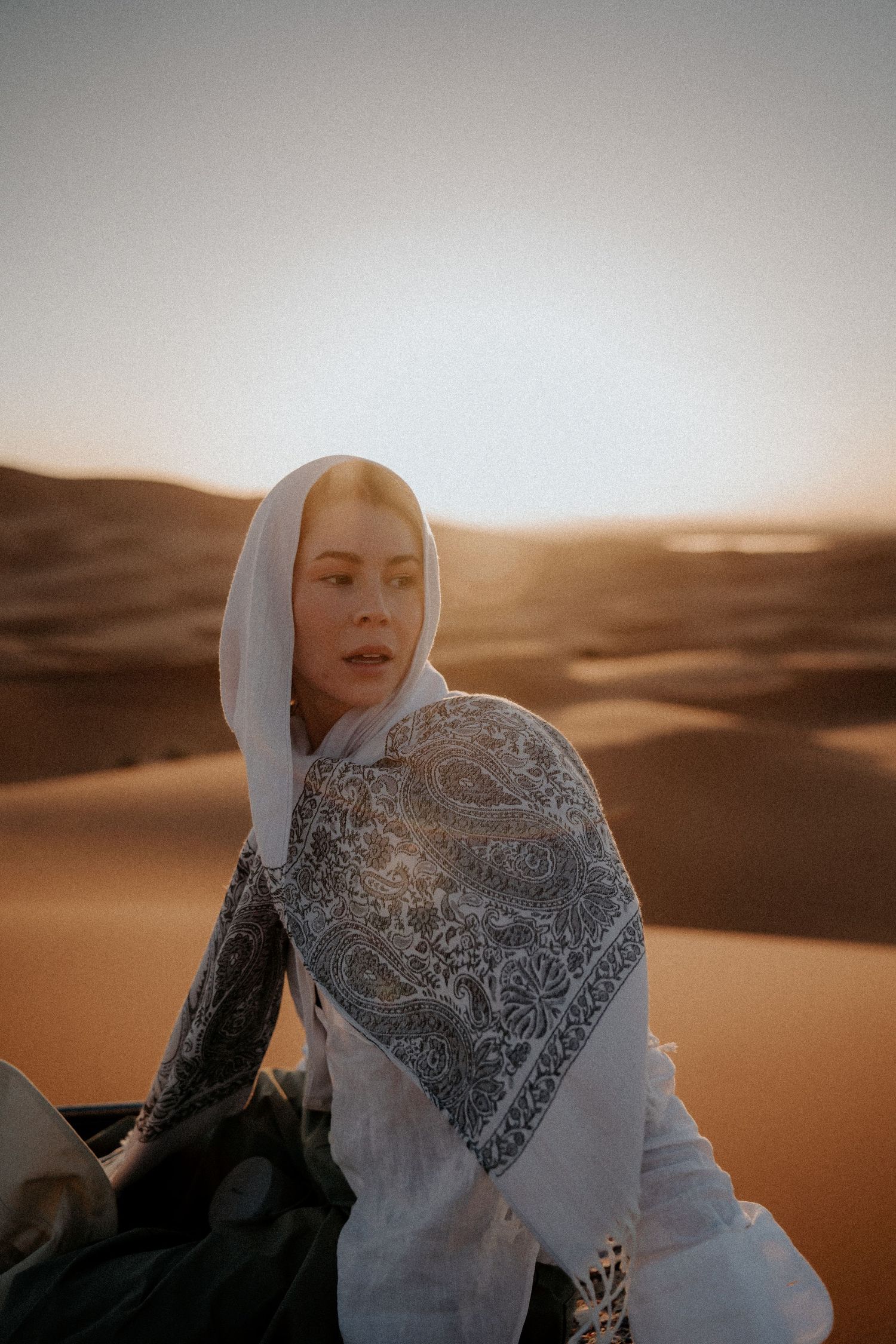 Figure in white headscarf and patterned dress amid desert dunes at sunset.