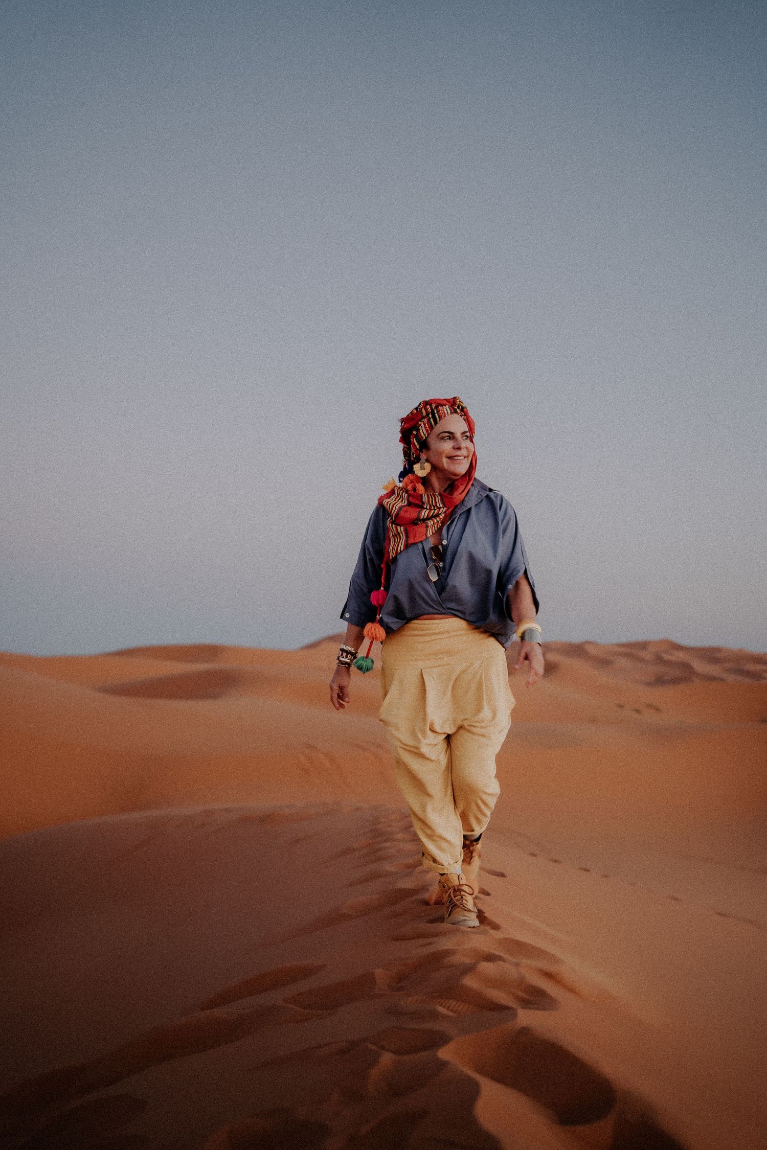A traveler walks along a sand dune in traditional desert attire at sunset.