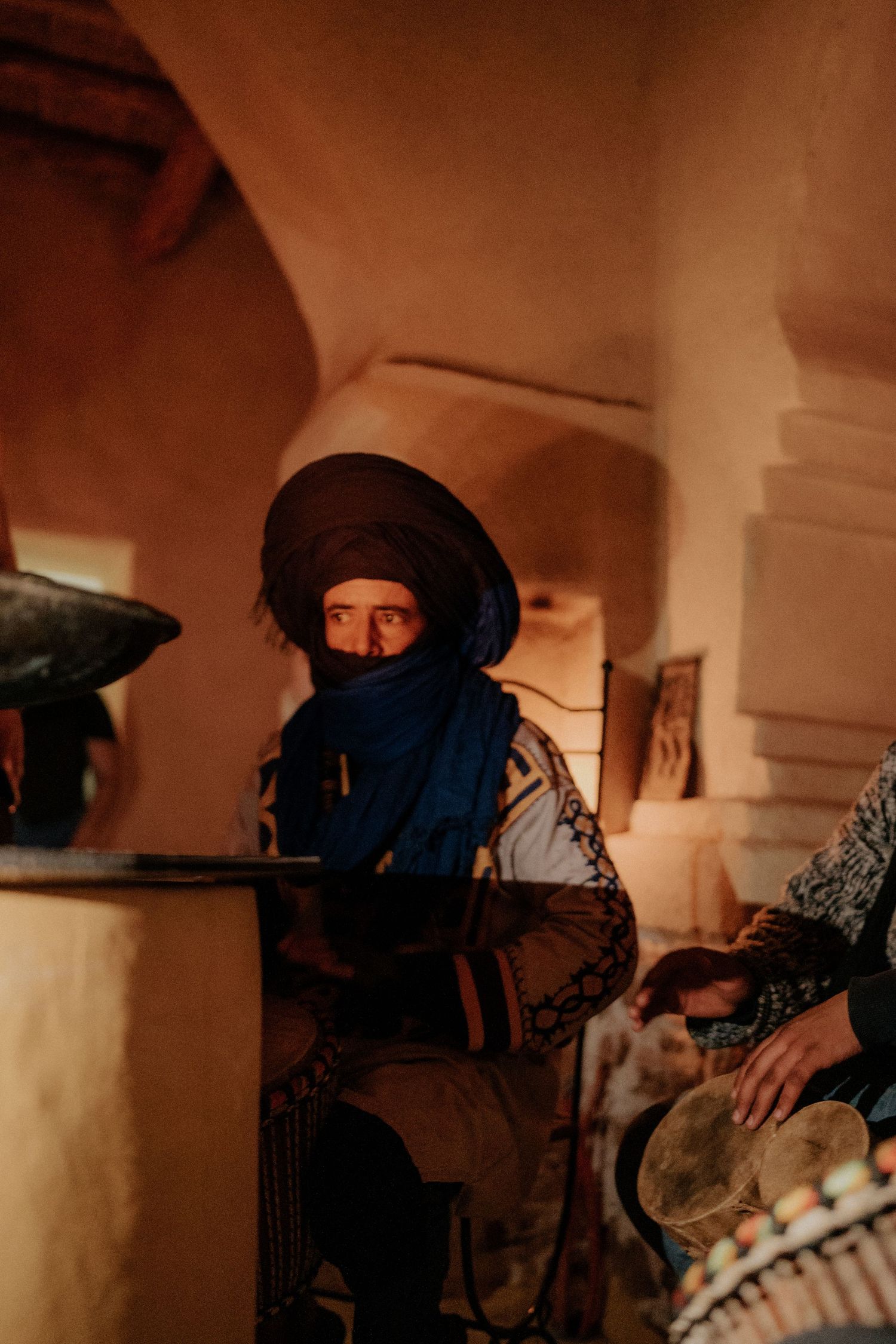 Traditional desert musician wearing blue head wrap plays instrument in warm candlelit setting.