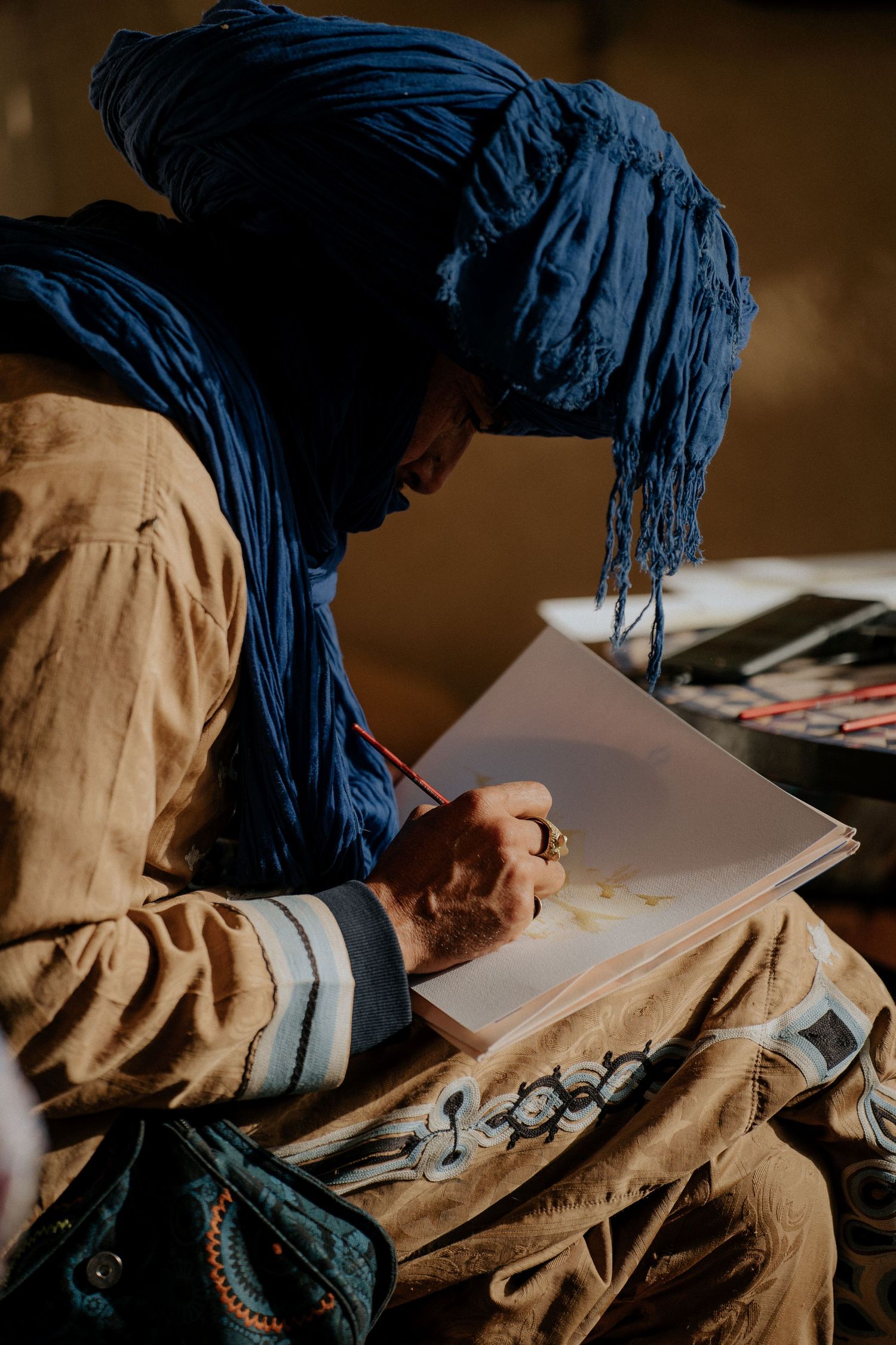 Person in blue traditional headwrap writing in notebook under natural lighting.