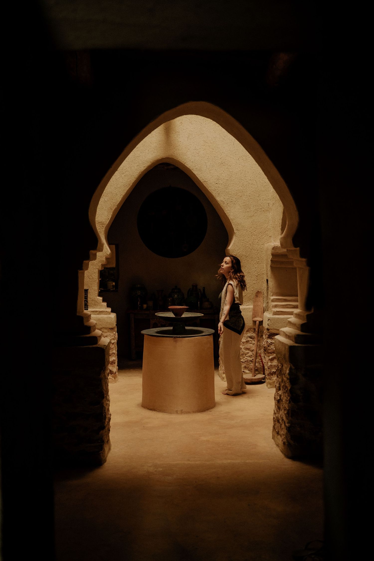 View through Moroccan archway of person standing by circular well in sepia-toned lighting.